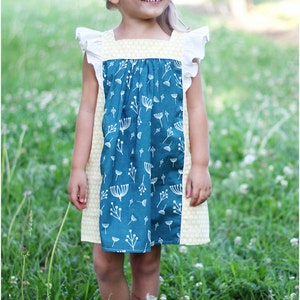 May include: A young girl wearing a blue and yellow dress with white floral print. The dress has a gathered bodice and ruffled sleeves. The girl is standing in a field of green grass.