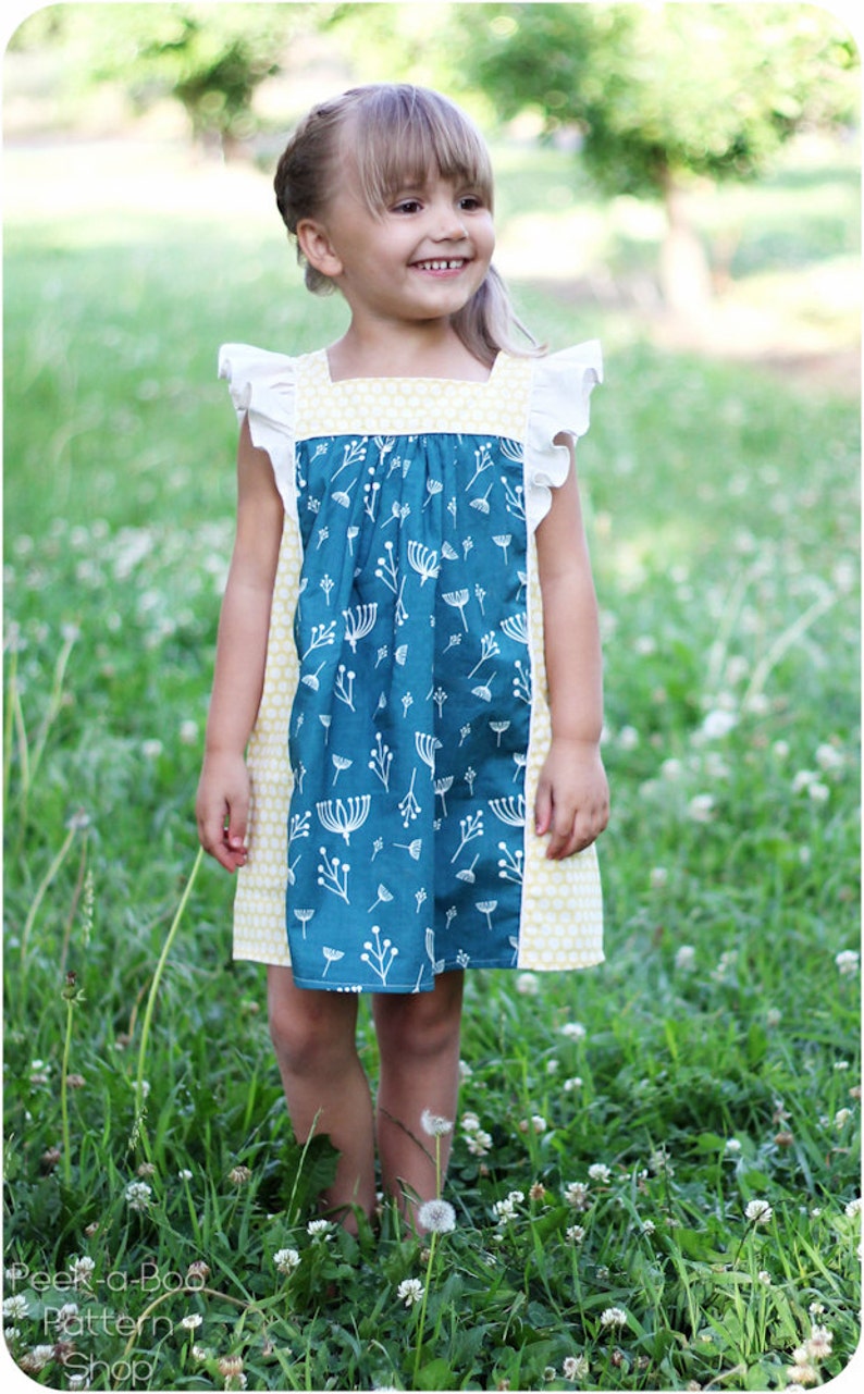 May include: A young girl wearing a blue and yellow dress with white floral print. The dress has a gathered bodice and ruffled sleeves. The girl is standing in a field of green grass.