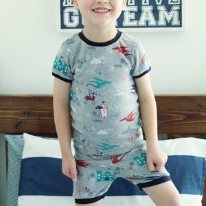 Puede incluir: Un ni&ntilde;o peque&ntilde;o con una camiseta de pijama de manga corta gris y blanca con ribete azul. La pijama tiene un estampado de castillos, dragones y caballeros. El ni&ntilde;o est&aacute; de pie frente a una cabecera de madera con una almohada a rayas azules y blancas.