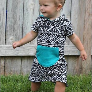 May include: A toddler wearing a black and white geometric print hooded dress with a turquoise pocket. The dress has short sleeves and a loose fit. The child is standing on green grass.