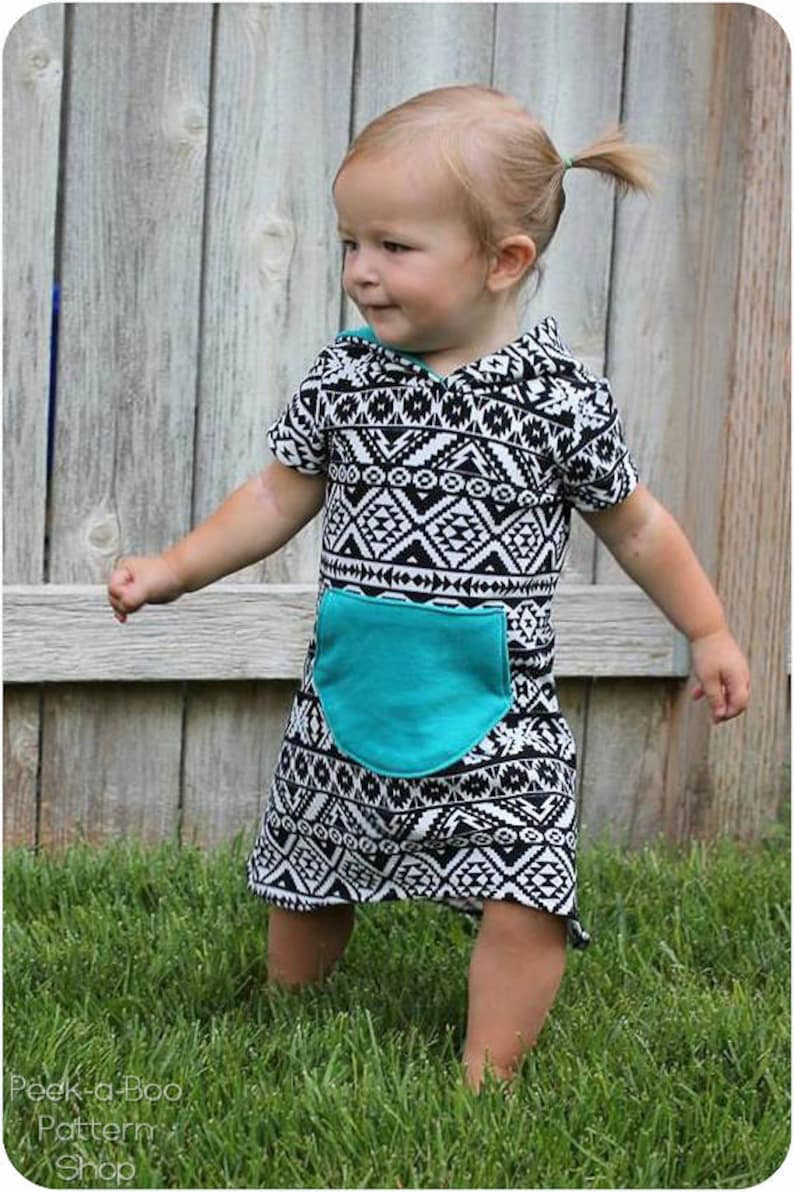 May include: A toddler wearing a black and white geometric print hooded dress with a turquoise pocket. The dress has short sleeves and a loose fit. The child is standing on green grass.