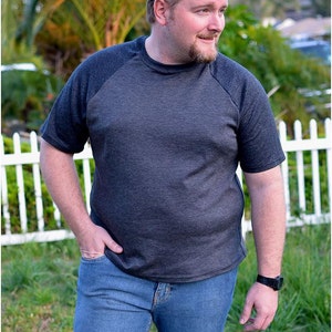 May include: A man wearing a dark gray and black raglan t-shirt with a crew neck and short sleeves. He is wearing blue jeans and a watch on his left wrist.
