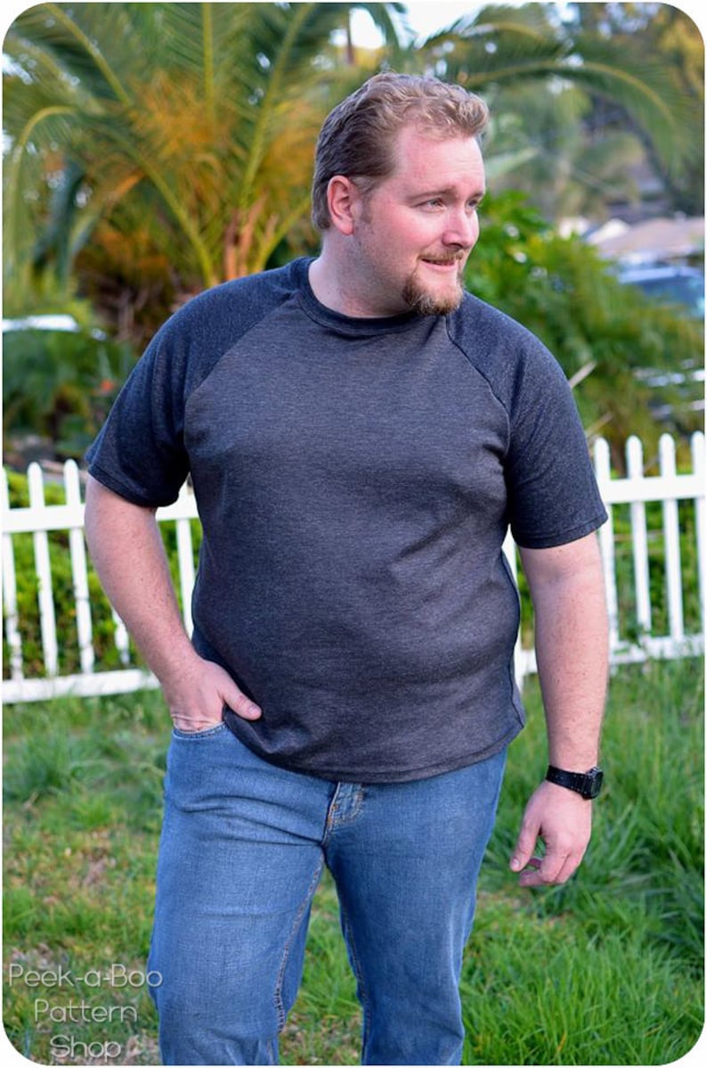 May include: A man wearing a dark gray and black raglan t-shirt with a crew neck and short sleeves. He is wearing blue jeans and a watch on his left wrist.