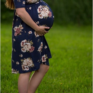 May include: A pregnant person wearing a navy blue floral maternity dress with a fitted waist. The dress has a short sleeve and a floral pattern with pink, white, and brown flowers. The person is standing in a grassy field.