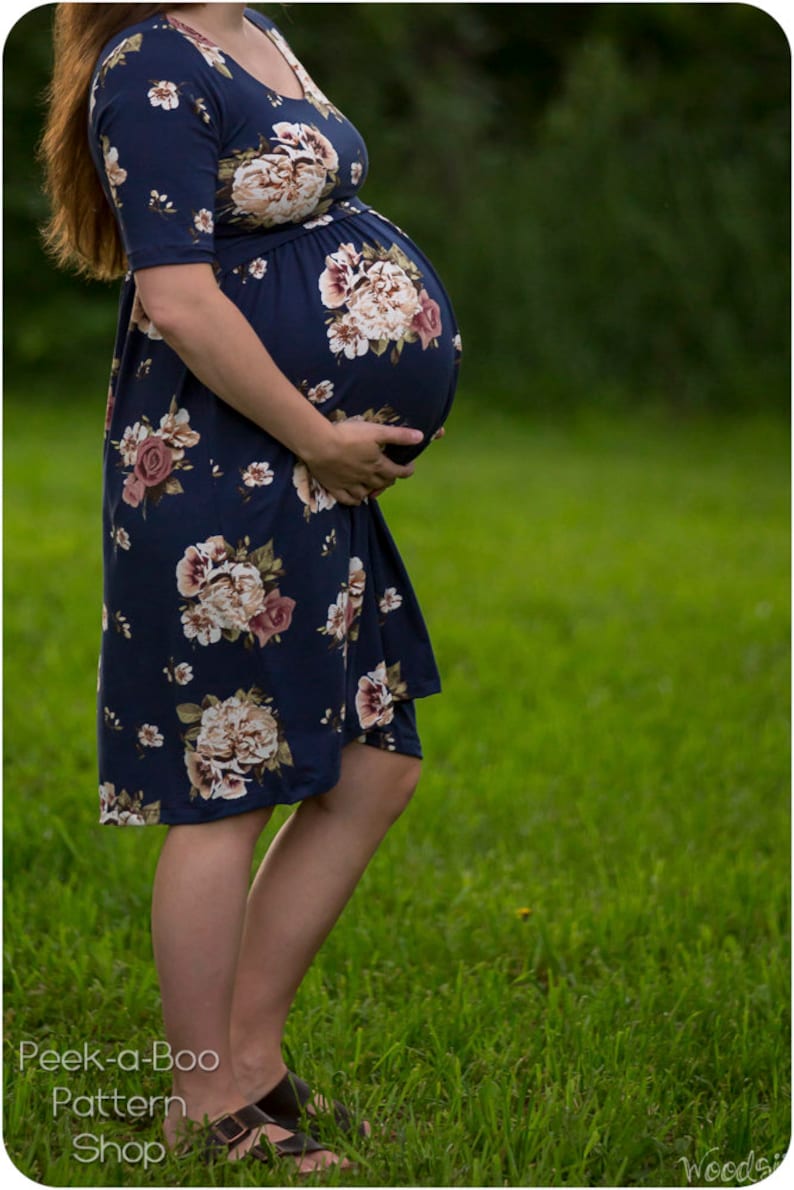 May include: A pregnant person wearing a navy blue floral maternity dress with a fitted waist. The dress has a short sleeve and a floral pattern with pink, white, and brown flowers. The person is standing in a grassy field.