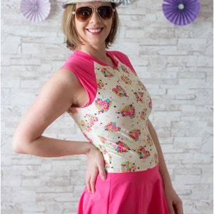 May include: A woman wearing a white and pink swimsuit with a floral print and a pink skirt. She is wearing a straw hat with a colorful band and sunglasses. The background is a white brick wall with paper fans.