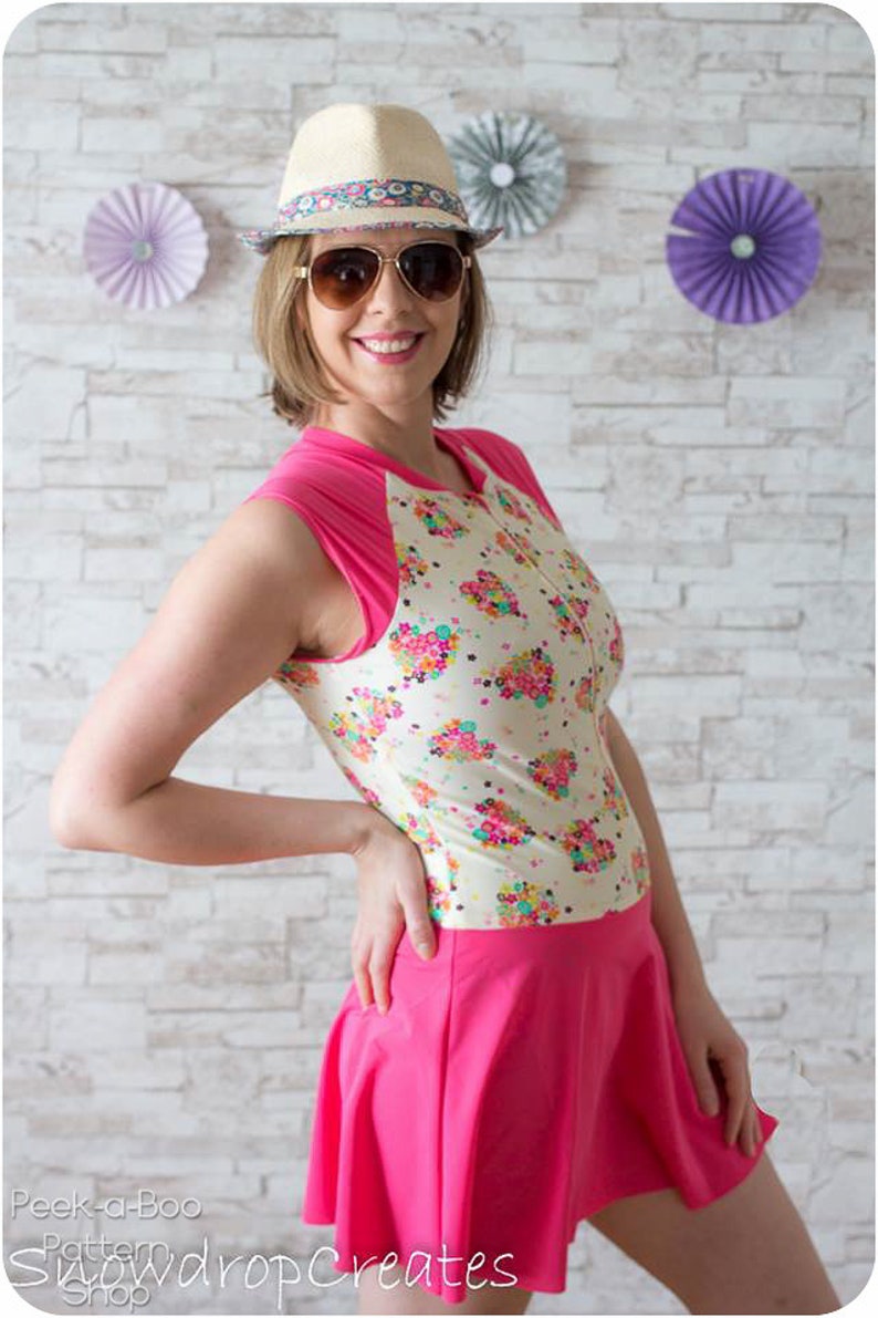 May include: A woman wearing a white and pink swimsuit with a floral print and a pink skirt. She is wearing a straw hat with a colorful band and sunglasses. The background is a white brick wall with paper fans.