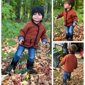 May include: A young child wearing a brown quilted jacket with black trim, blue jeans, and black boots. The child is standing in a pile of autumn leaves.