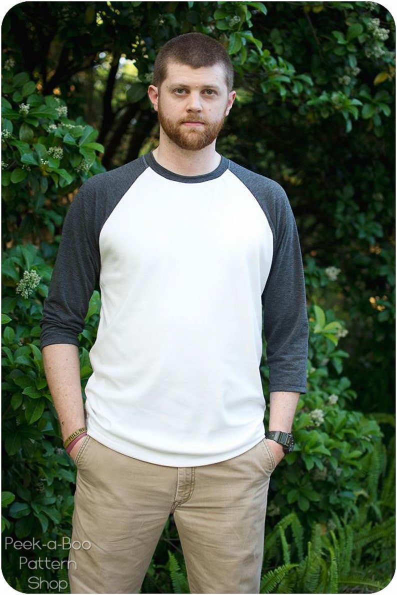 May include: A man wearing a white and gray baseball tee shirt with a gray and white color block design. He is standing in front of a green leafy background.