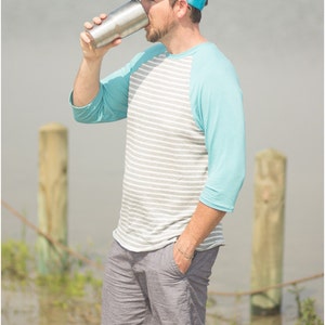May include: A man wearing a blue and gray baseball cap, a striped gray and white shirt with a blue sleeve, and gray shorts, is drinking from a stainless steel tumbler.
