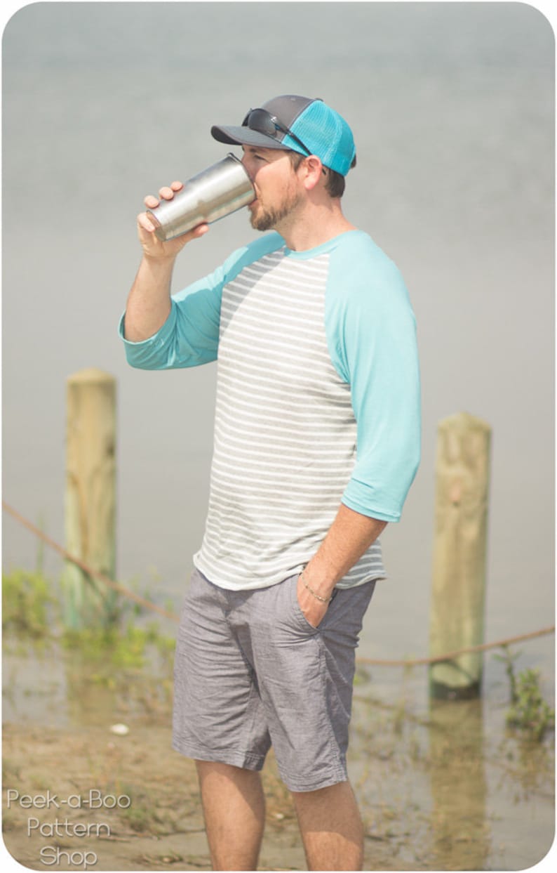 May include: A man wearing a blue and gray baseball cap, a striped gray and white shirt with a blue sleeve, and gray shorts, is drinking from a stainless steel tumbler.