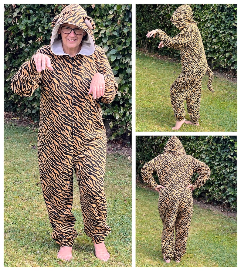 May include: A person wearing a tiger-print onesie with a hood. The onesie has a tail and is brown and black. The person is standing in a grassy area.