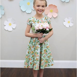 May include: A young girl wearing a light blue floral dress with a pink sash and white sandals. She is holding a bouquet of pink flowers and is standing in front of a wall decorated with paper flowers.