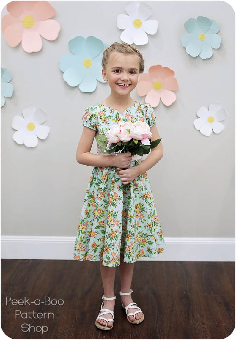May include: A young girl wearing a light blue floral dress with a pink sash and white sandals. She is holding a bouquet of pink flowers and is standing in front of a wall decorated with paper flowers.