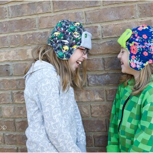May include: Two children wearing colorful patterned winter hats with ear flaps. The girl on the left is wearing a gray hooded sweatshirt with a white pattern. The girl on the right is wearing a green and black checkered hooded sweatshirt.  Peek-a-Boo Pattern Shop.