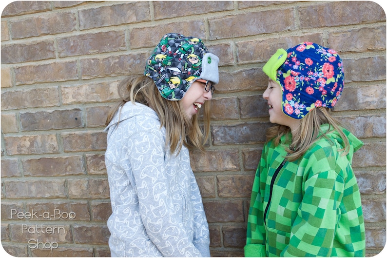 May include: Two children wearing colorful patterned winter hats with ear flaps. The girl on the left is wearing a gray hooded sweatshirt with a white pattern. The girl on the right is wearing a green and black checkered hooded sweatshirt.  Peek-a-Boo Pattern Shop.