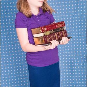 Puede incluir: Una persona joven con una camisa polo morada y una falda azul marino sostiene una pila de libros en sus brazos. Los libros tienen cubiertas rojas, doradas y negras. La persona est&aacute; de pie frente a un fondo azul con lunares blancos.