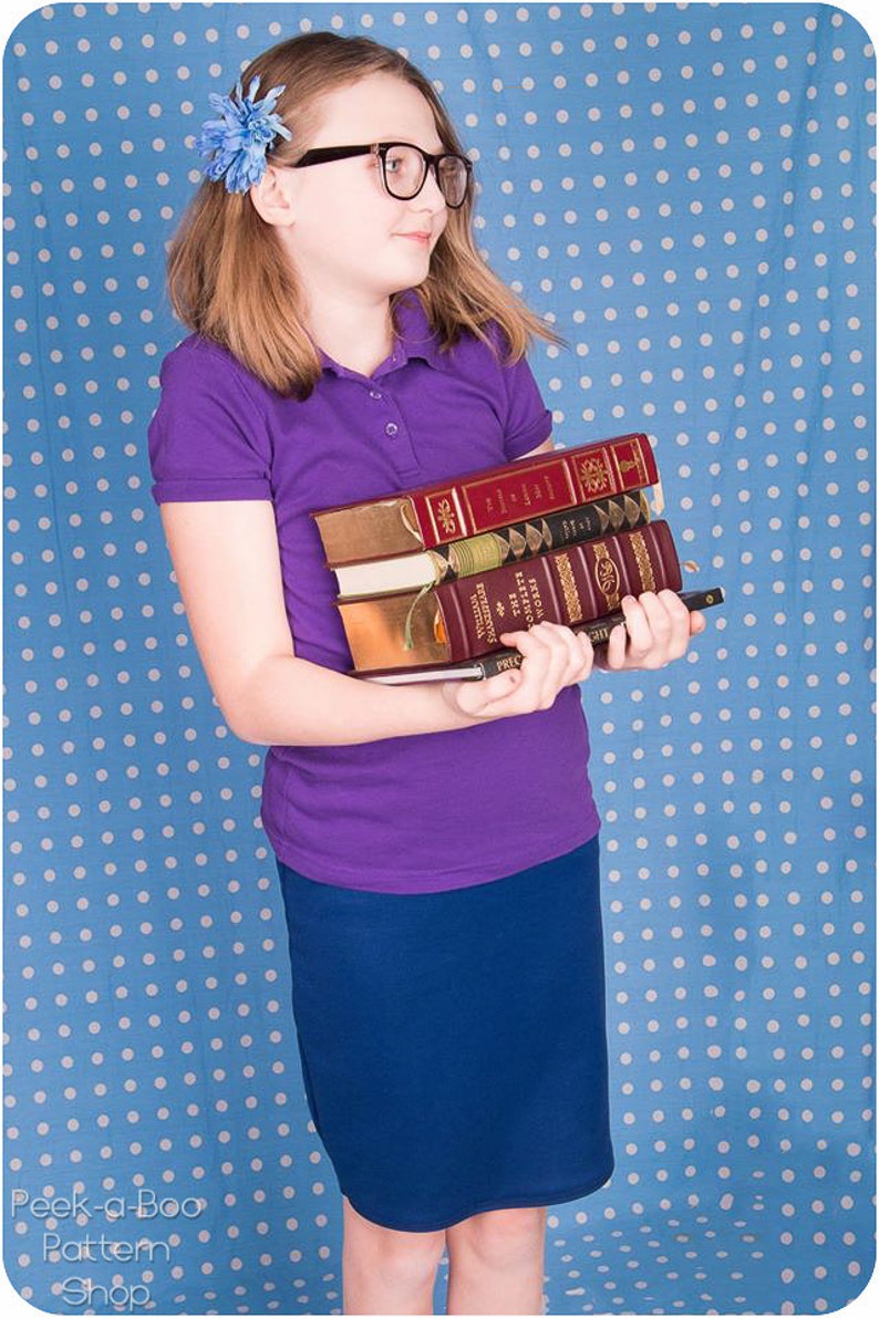 Puede incluir: Una persona joven con una camisa polo morada y una falda azul marino sostiene una pila de libros en sus brazos. Los libros tienen cubiertas rojas, doradas y negras. La persona est&aacute; de pie frente a un fondo azul con lunares blancos.