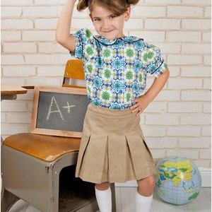 May include: A young girl wearing a white and blue patterned blouse, a khaki pleated skirt, white socks, and blue and white saddle shoes. She is holding a red apple and a book on her head. The book has the text "Prec 2 Good Stories". There is a chalkboard behind her with the text "A+" written on it.