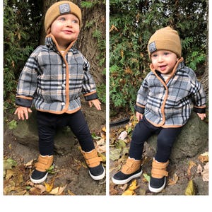 May include: Two toddlers wearing brown and black checked jackets with brown trim, brown knitted hats with a black logo, and black and brown boots. The toddlers are sitting on rocks in a woodland setting.