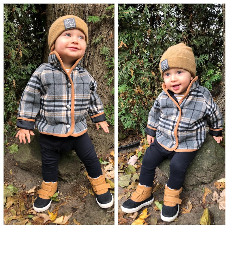 May include: Two toddlers wearing brown and black checked jackets with brown trim, brown knitted hats with a black logo, and black and brown boots. The toddlers are sitting on rocks in a woodland setting.