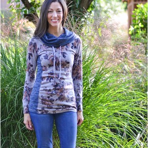 May include: A woman wearing a long-sleeved floral print top with a cowl neck and a blue and gray color block design. She is wearing blue jeans and brown boots. The top has a drawstring at the neck. The image is taken outdoors in front of a green background.