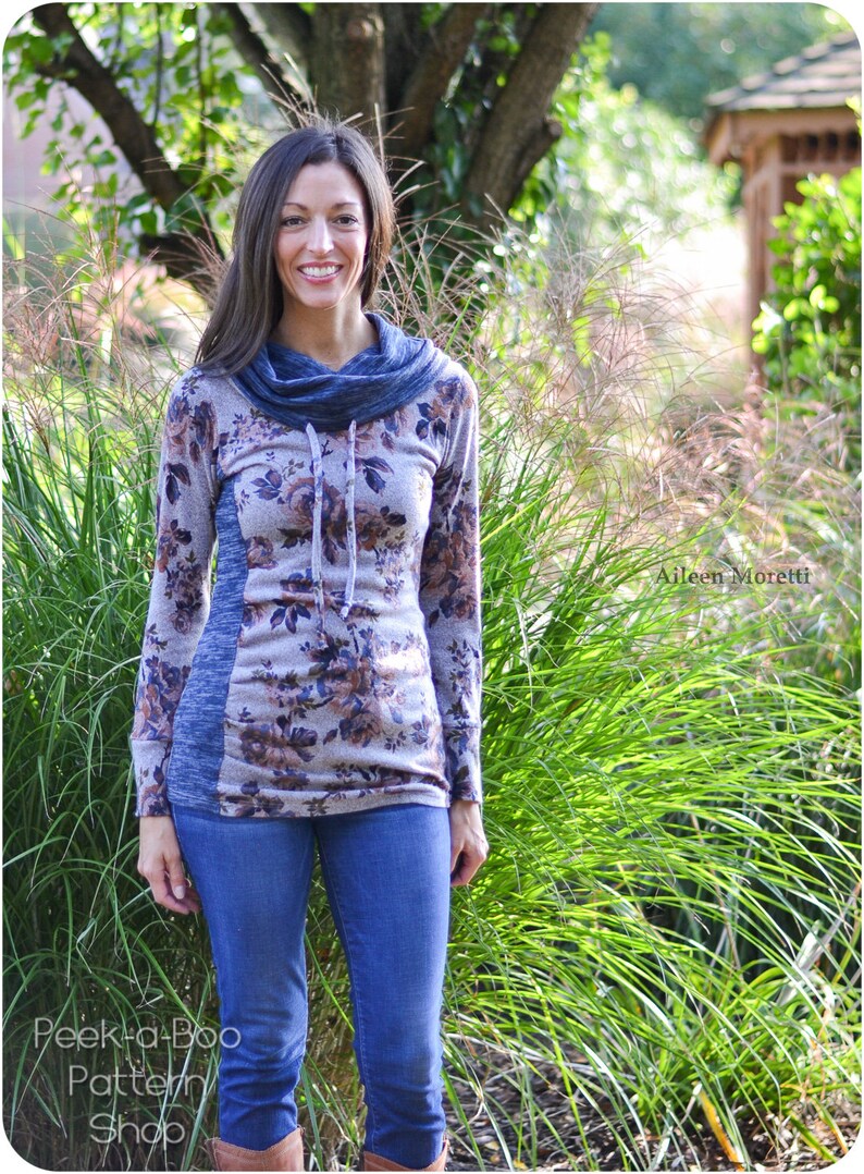 May include: A woman wearing a long-sleeved floral print top with a cowl neck and a blue and gray color block design. She is wearing blue jeans and brown boots. The top has a drawstring at the neck. The image is taken outdoors in front of a green background.