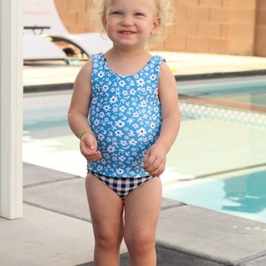 May include: A young girl wearing a blue and white floral swimsuit with a black and white checkered bottom. She is standing in front of a swimming pool.