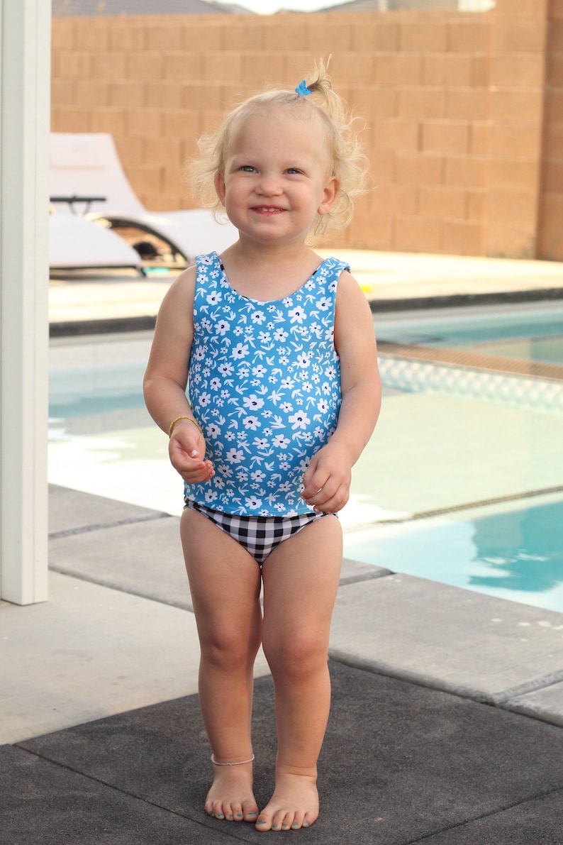 May include: A young girl wearing a blue and white floral swimsuit with a black and white checkered bottom. She is standing in front of a swimming pool.