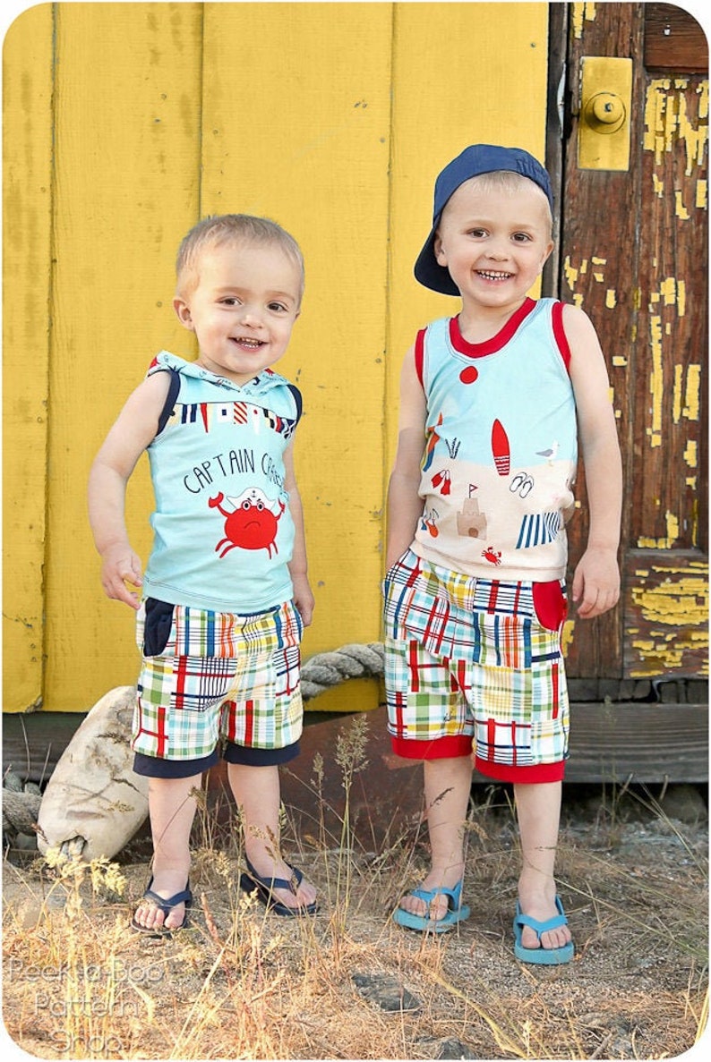May include: Two young boys wearing summer clothing. The boy on the left is wearing a blue sleeveless shirt with a red crab graphic and the text "Captain Crew". He is also wearing plaid shorts. The boy on the right is wearing a blue sleeveless shirt with a beach scene graphic and plaid shorts. Both boys are wearing blue flip-flops.