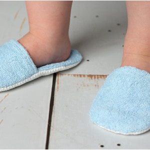 K&ouml;nnte beinhalten: Ein Paar hellblaue Frottee-Babyschuhe mit wei&szlig;em Besatz. Die Schuhe liegen auf einem wei&szlig;en Holzboden.