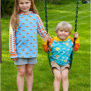 May include: Two children, a girl and a boy, are playing on a swing set. The girl is wearing a blue and orange patterned long-sleeved shirt and gray shorts. The boy is wearing a blue and orange patterned long-sleeved shirt with a hot air balloon design and khaki shorts. The swing set is green and has a chain.
