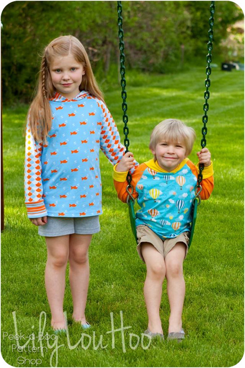 May include: Two children, a girl and a boy, are playing on a swing set. The girl is wearing a blue and orange patterned long-sleeved shirt and gray shorts. The boy is wearing a blue and orange patterned long-sleeved shirt with a hot air balloon design and khaki shorts. The swing set is green and has a chain.