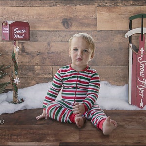 May include: A young child wearing red and green striped pajamas sits on a wood floor in front of a wood wall. The child is sitting in front of a small Christmas tree and a red sled with the words "Snow Flyer" written on it. There is a red mailbox with the words "Santa Mail" on it.