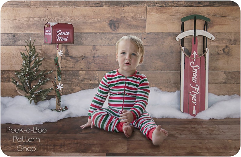 May include: A young child wearing red and green striped pajamas sits on a wood floor in front of a wood wall. The child is sitting in front of a small Christmas tree and a red sled with the words "Snow Flyer" written on it. There is a red mailbox with the words "Santa Mail" on it.