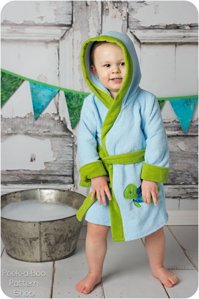 May include: A light blue hooded robe with green trim and a green belt. The robe has a small embroidered turtle on the front. A young child is wearing the robe.