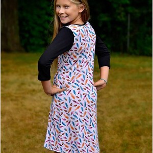 May include: A young girl wearing a white dress with a colorful feather print. The dress has a black long-sleeved shirt underneath. The girl is standing in a grassy area.