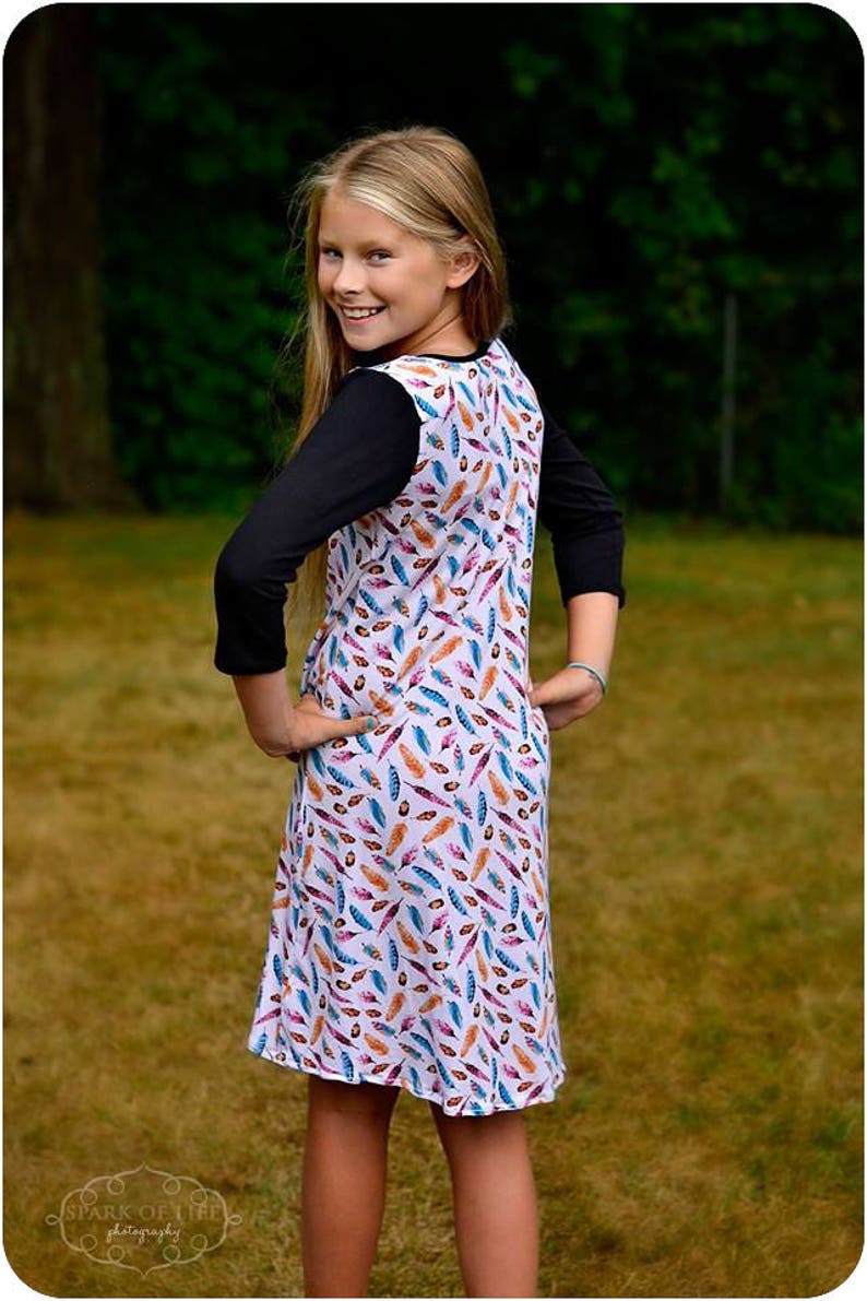 May include: A young girl wearing a white dress with a colorful feather print. The dress has a black long-sleeved shirt underneath. The girl is standing in a grassy area.