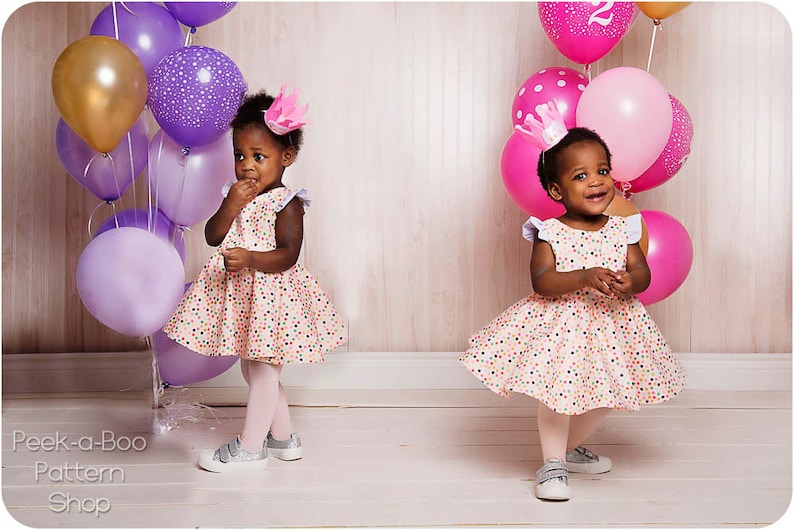 May include: Two young girls wearing pink and white dresses with colorful polka dots and crowns on their heads. They are surrounded by purple and pink balloons.