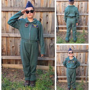May include: A child wearing a green flight suit with a blue pilot's cap, sunglasses, and patches. The suit has a zipper and a drawstring waist. The patches include a squadron emblem and the name "J. POE".