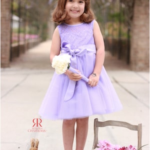 May include: A young girl in a lavender dress with a white lace bodice and a satin bow holds a bouquet of white flowers. She is standing in front of a wooden crate filled with pink and purple flowers. The crate has a sign that reads "Flowers & Garden".
