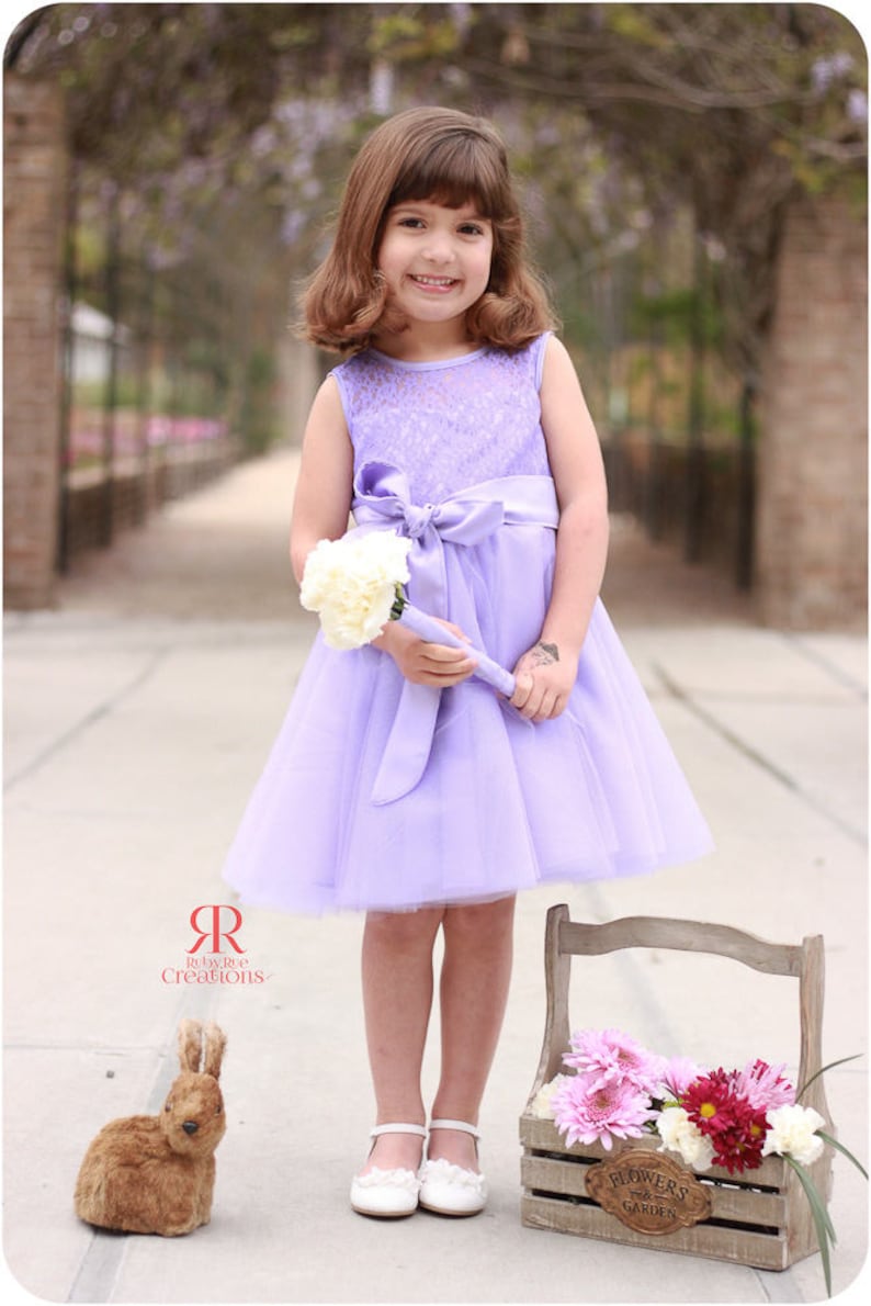 May include: A young girl in a lavender dress with a white lace bodice and a satin bow holds a bouquet of white flowers. She is standing in front of a wooden crate filled with pink and purple flowers. The crate has a sign that reads "Flowers & Garden".