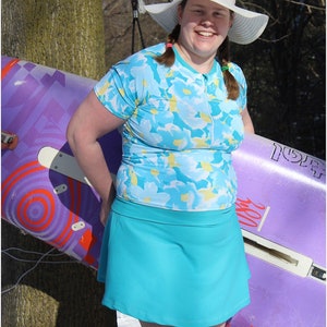 May include: A woman wearing a white sun hat, a blue and white floral print short-sleeved shirt, and a turquoise skirt. She is holding a purple and orange surfboard.