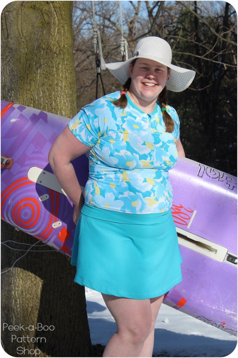 May include: A woman wearing a white sun hat, a blue and white floral print short-sleeved shirt, and a turquoise skirt. She is holding a purple and orange surfboard.