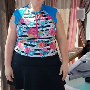May include: A woman wearing a black and white striped swimsuit with a floral print and a black skirt. The swimsuit has a blue zipper and blue sleeves. The woman is standing in front of a hot tub.