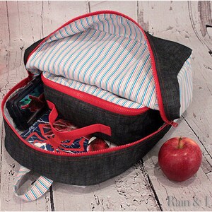 May include: A denim backpack with a red zipper and a striped lining. The backpack is open and has a lunch bag inside. There is a red apple on the table next to the backpack.