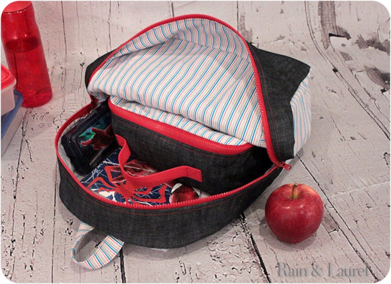May include: A denim backpack with a red zipper and a striped lining. The backpack is open and has a lunch bag inside. There is a red apple on the table next to the backpack.