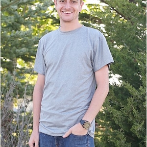 May include: A man wearing a gray t-shirt and blue jeans. He is standing in front of a green bush and smiling.