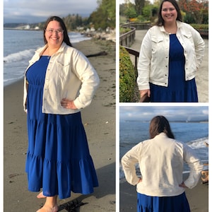 May include: A cream-colored denim jacket with button closures and pockets. The jacket is worn over a long, tiered, royal blue dress. The dress has a textured pattern. The background shows a beach scene with water and trees.