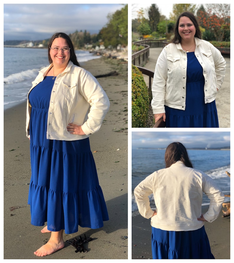 May include: A cream-colored denim jacket with button closures and pockets. The jacket is worn over a long, tiered, royal blue dress. The dress has a textured pattern. The background shows a beach scene with water and trees.